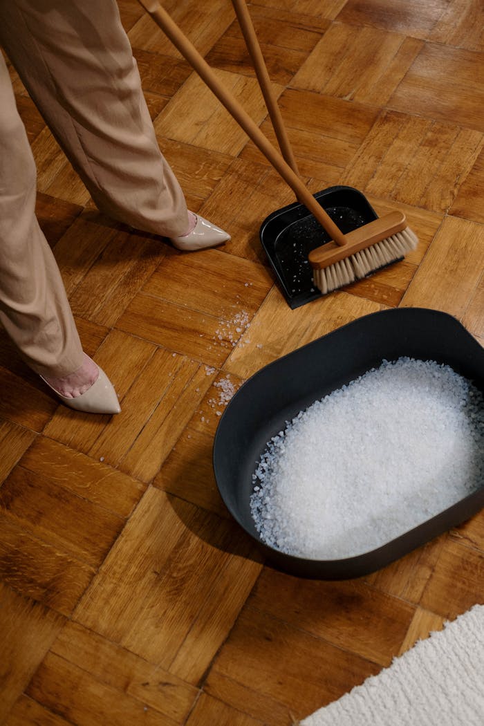 A woman sweeping salt with a broom onto a dustpan on a parquet floor indoors.